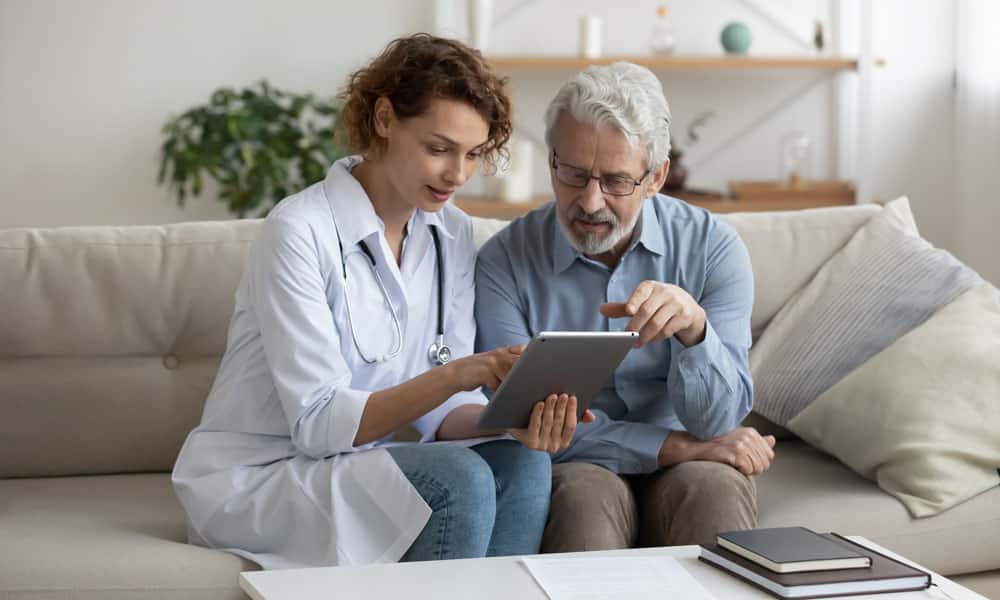 A female doctor in a white coat and with a stethoscope around her neck sits on a couch next to an older male patient. She is holding a tablet and pointing to the screen while he also points, and they both look at it intently.
