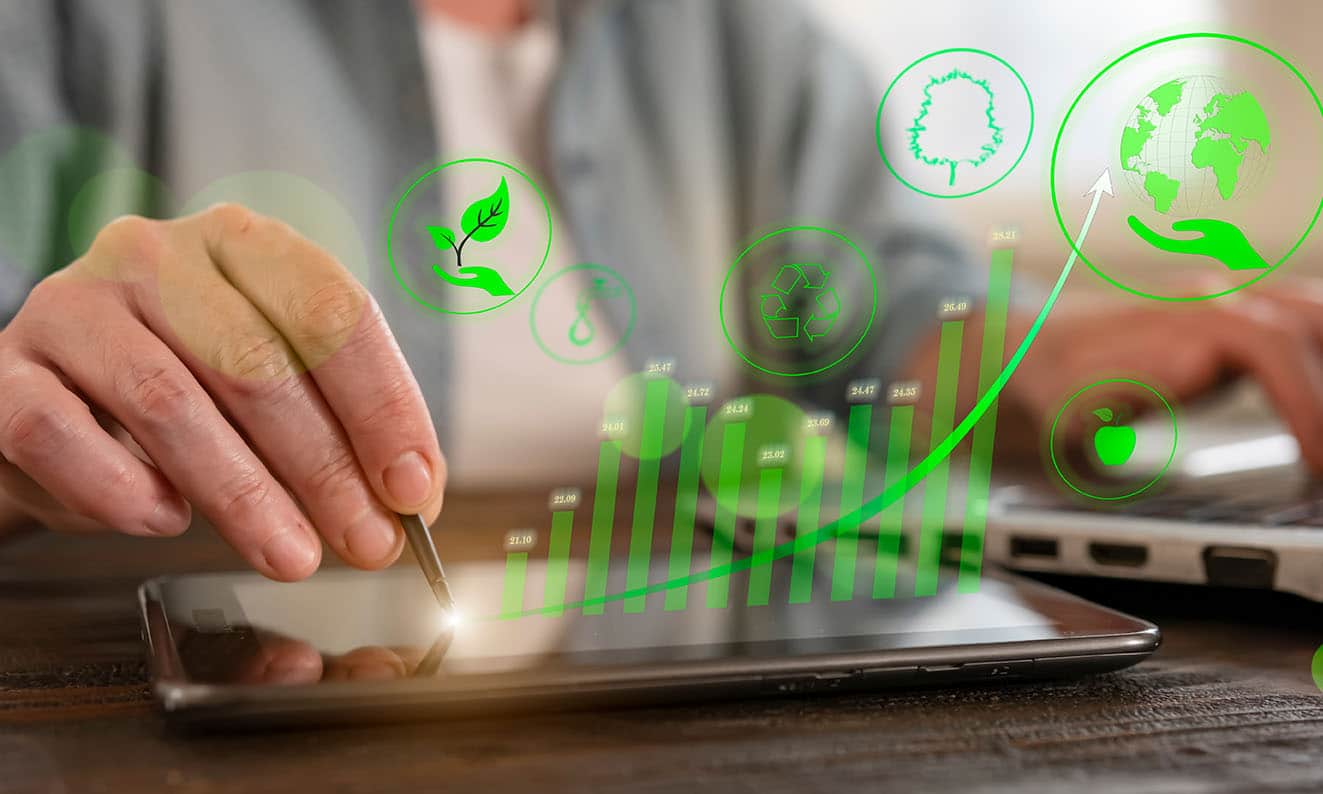 A person's hands interact with a tablet displaying a rising green line graph superimposed over icons related to sustainability, including a recycling symbol, a tree, a leaf, and a globe with a hand